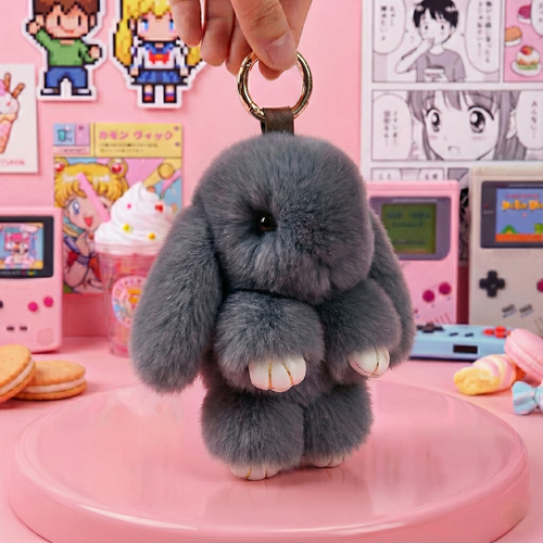 Gray fluffy bunny keychain held by a hand against a colorful background with anime posters and toys.